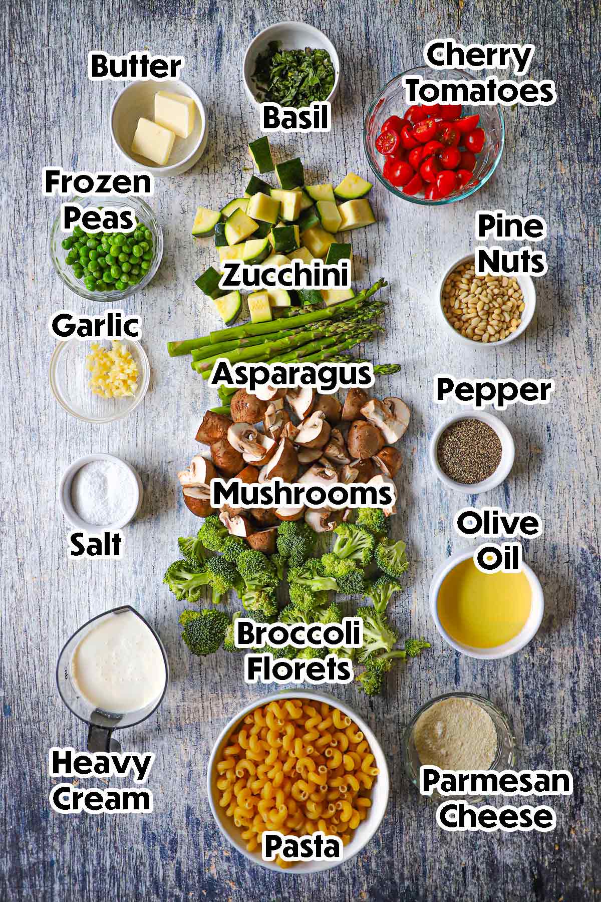 An arrangement of ingredients for Homemade Pasta Primavera on a grey wooden background including pasta, broccoli, mushrooms, asparagus, zucchini, cherry tomatoes, peas, garlic, cream, Parmesan, pine nuts, and seasonings.