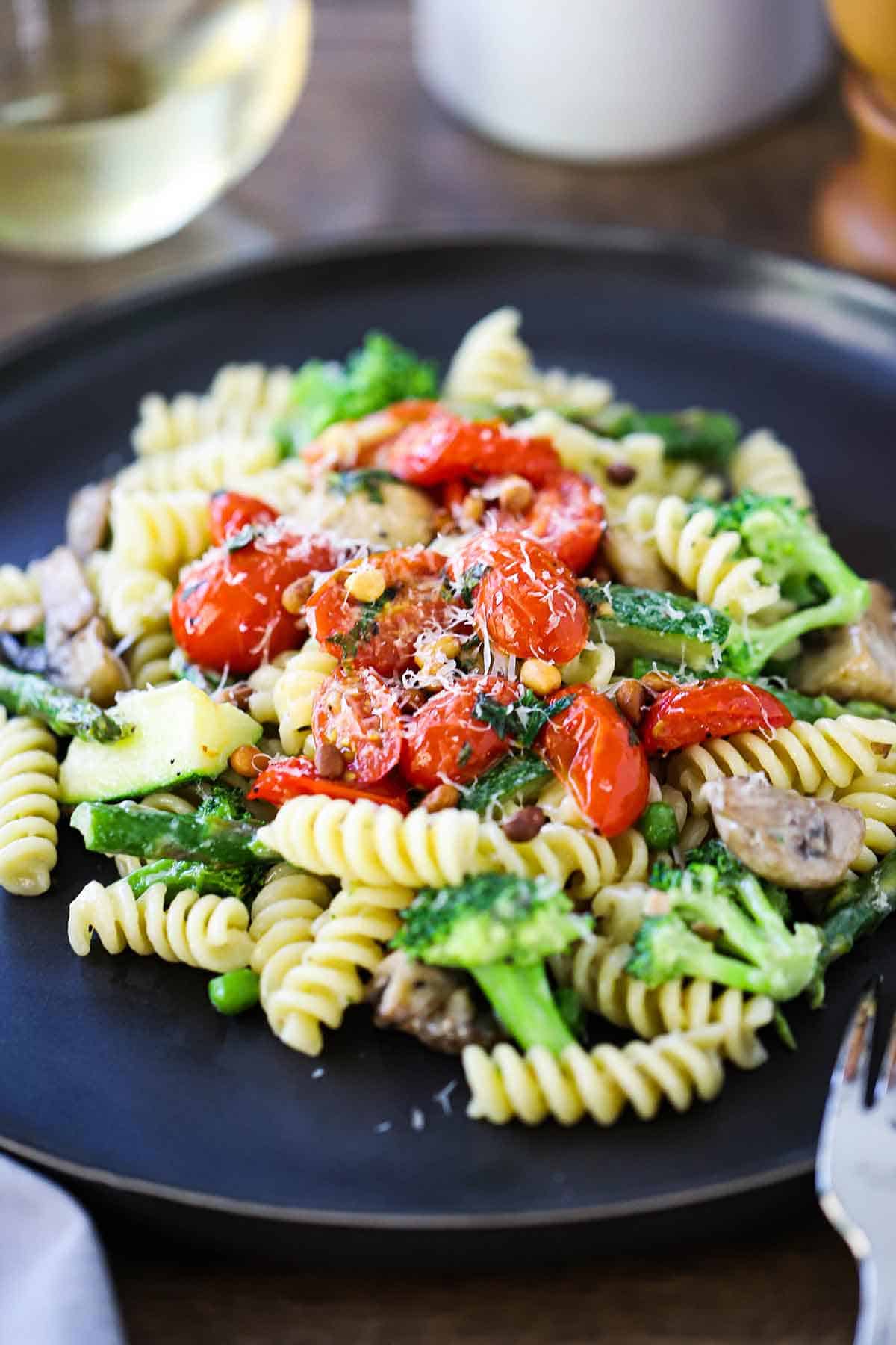 A straight-on view of a black dinner plate filled with a serving of homemade pasta primavera topped with sautéed cherry tomatoes, fresh basil, and grated Parmesan cheese.