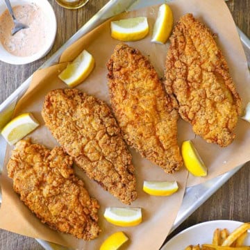 An overhead view of a large baking pan lined with brown paper and topped with four Southern fried catfish filets with lemon wedges around the sides of the pan.