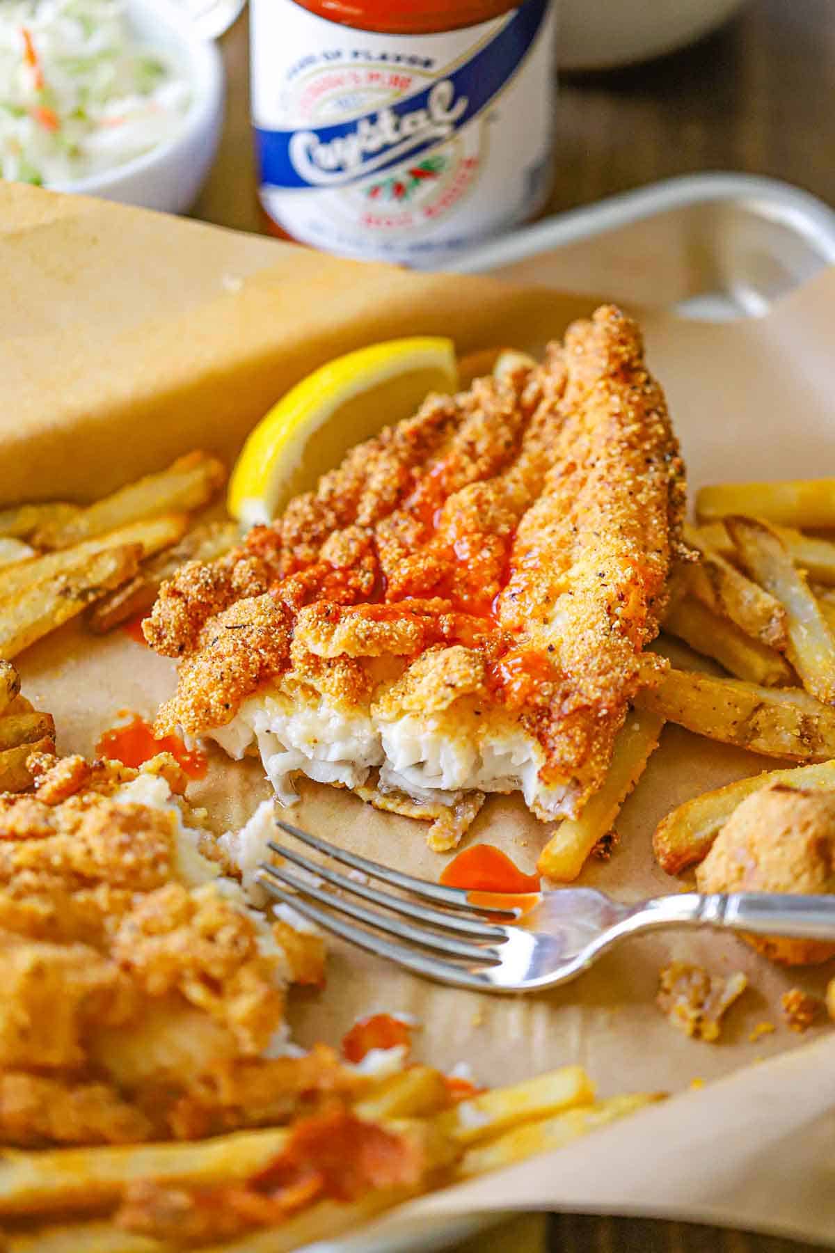 A straight-on view of a partially east Southern Fried Catfish filet that is topped with Crystal's Hot Sauce and has French fries next to the filet.