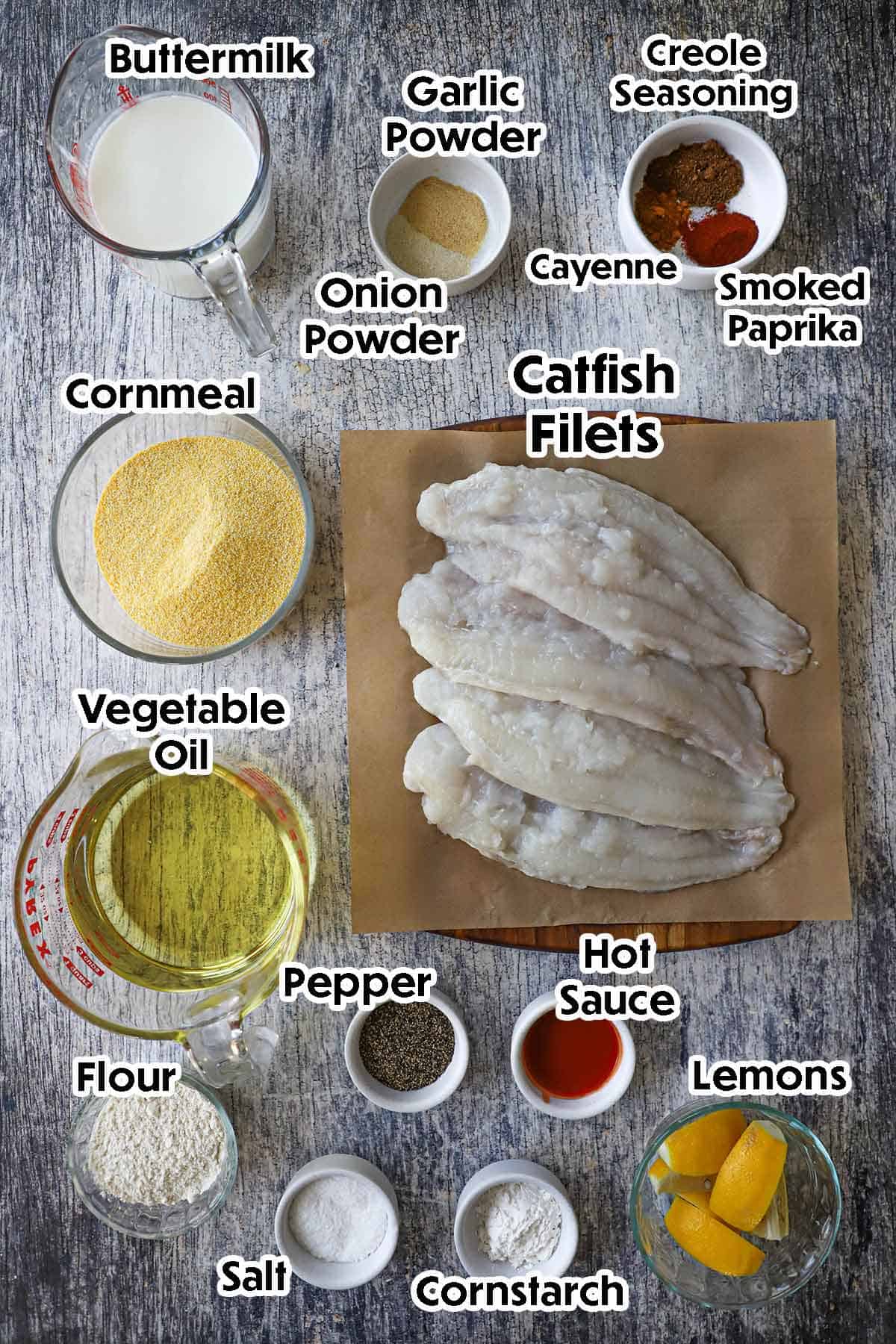 An arrangement of ingredients for Southern Fried Catfish on a grey wooden background including catfish filets, cornmeal, buttermilk, flour, oil, hot sauces, lemon wedges, and seasonings.
