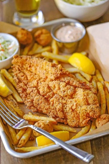 A close-up view of a Southern Fried Catfish on a small metal pan lined with brown paper along with French fries, a small bowl of coleslaw, and a small condiment holder containing Cajun remoulade.