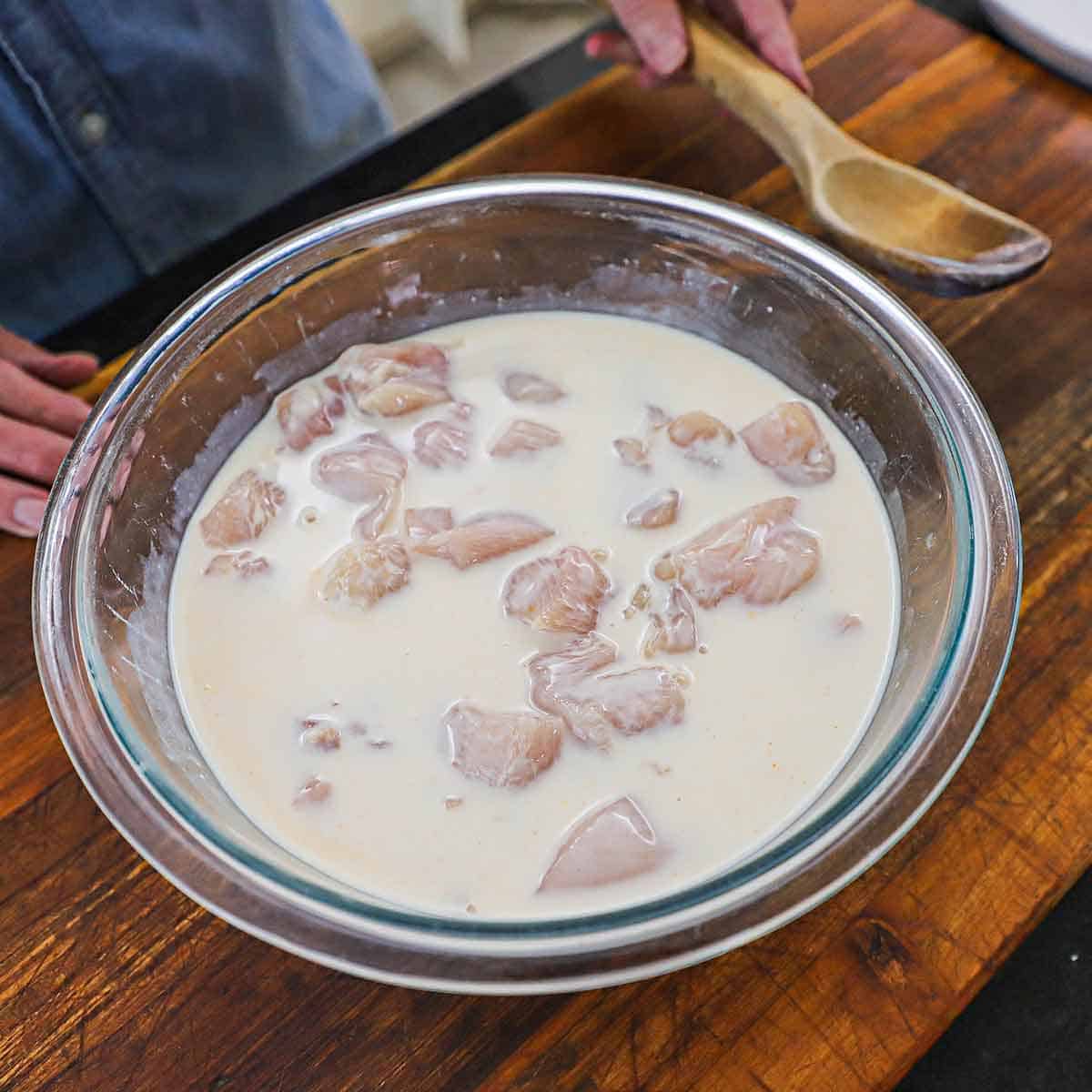 A person using a wooden spoon to submerge chunks of skinless chicken breasts in a buttermilk, pickle brine, and hot sauce mixture all in a large glass bowl.