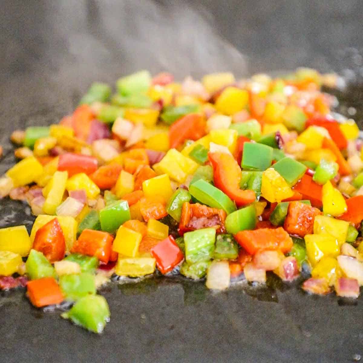 A close-up view of chopped red, yellow, and green bell peppers, along with chopped onion and jalapeño being sautéed in oil and butter on a griddle.