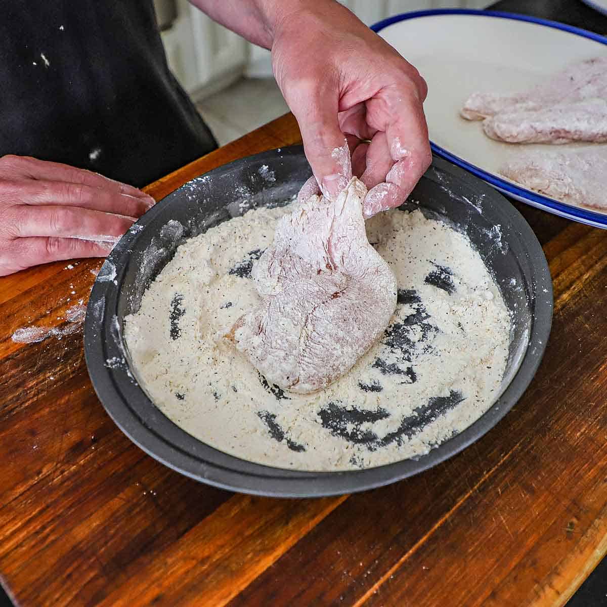 A person dredging a boneless, skinless chicken breast through seasoned flour in a round baking pan.