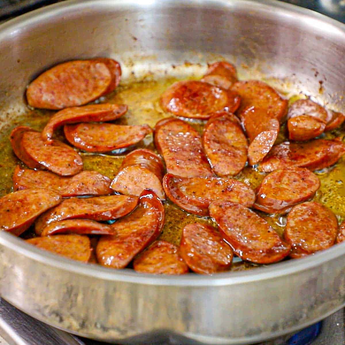 A large stainless steel skillet filled with sliced andouille sausage that has been seared in a olive oil on a gas stove.
