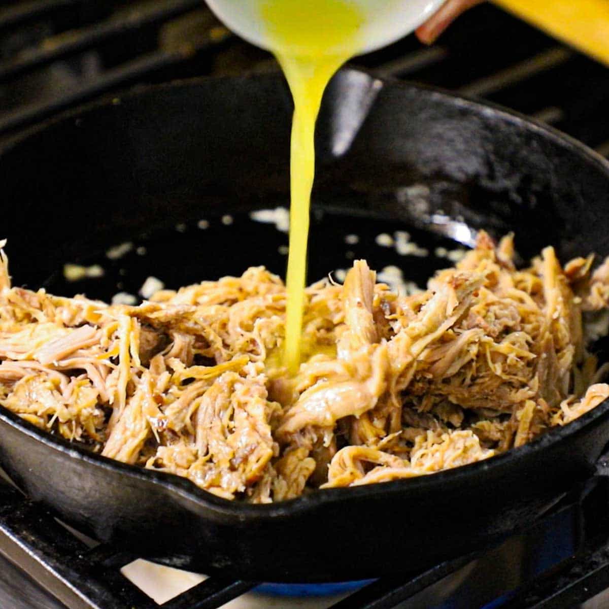 A person pouring orange juice from a small white bowl into a skillet that is filled with shredded pulled pork.