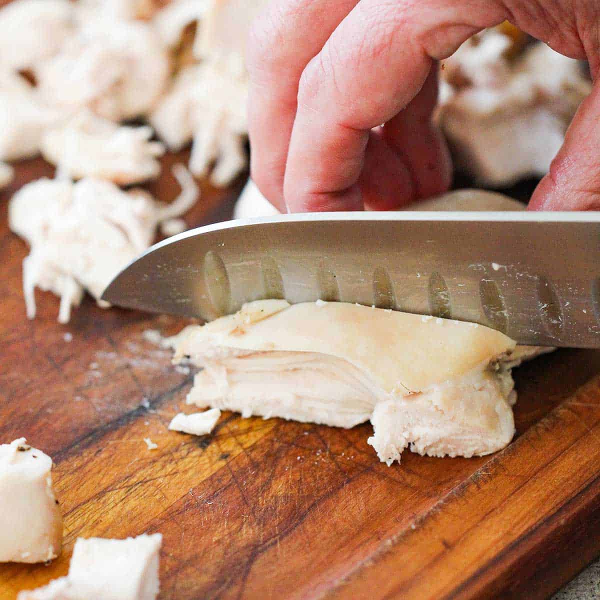 A person using a chef's knife to roughly chop a fully cooked chicken breast on a wooden cutting board.