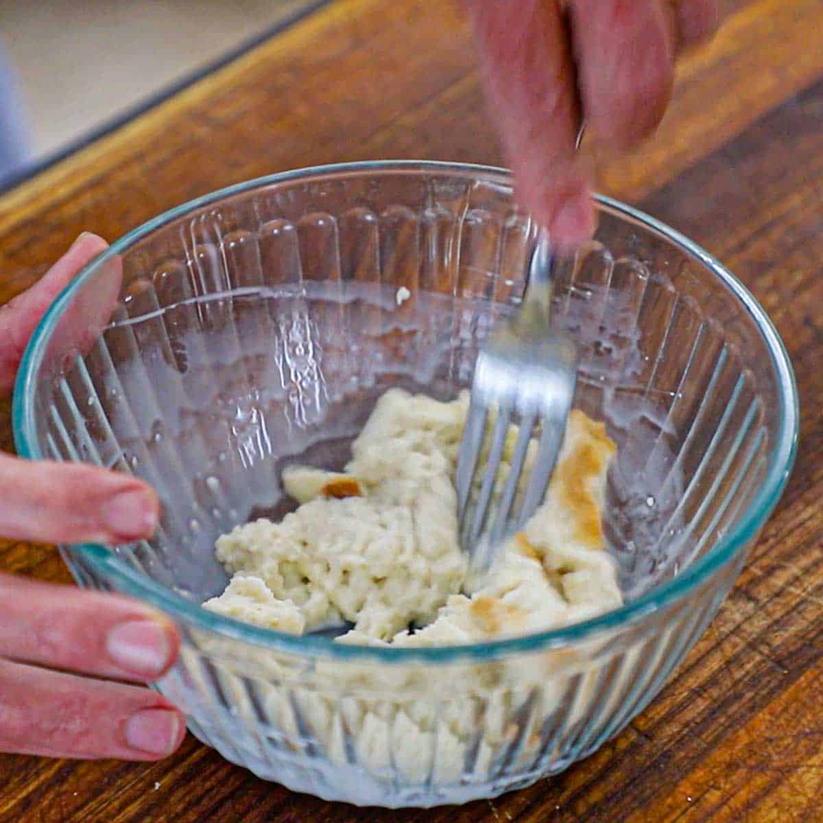 A person using a fork to mash white bread that has been soaked in whole milk in a small glass bowl on a wooden cutting board.