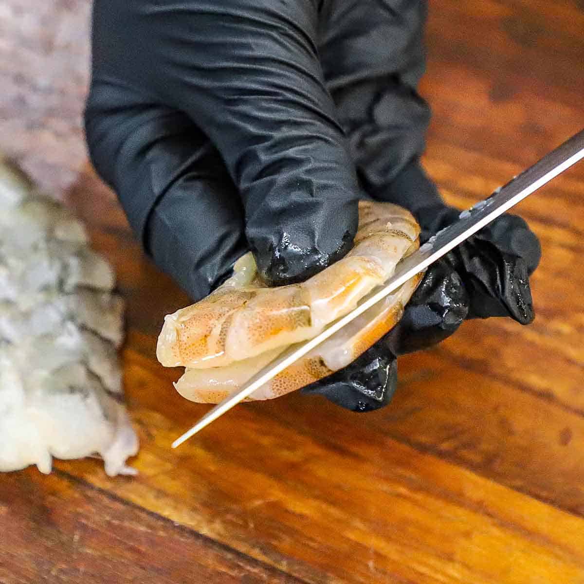 A person wearing black kitchen gloves holding an extra-large raw shrimp with the shell removed in one hand and using a paring knife in the other hand to cut a deep slit through the backside of the shrimp.