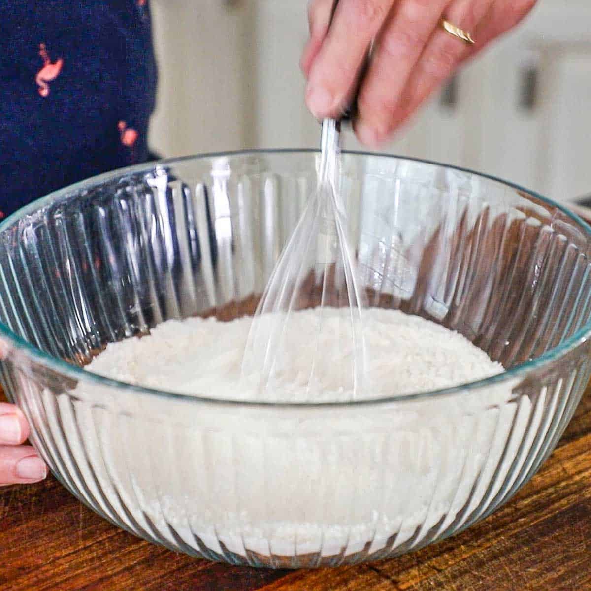 A person using a wire whisk to combine all-purpose with baking powder, baking soda, and salt in a large glass bowl on a wooden cutting board.