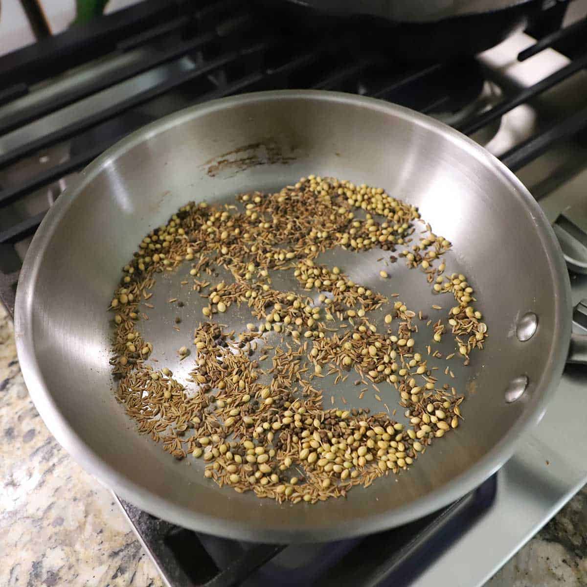 A silver skillet on a gas stove filled with coriander seeds, cumin seeds, and cardamom seeds being toasted.