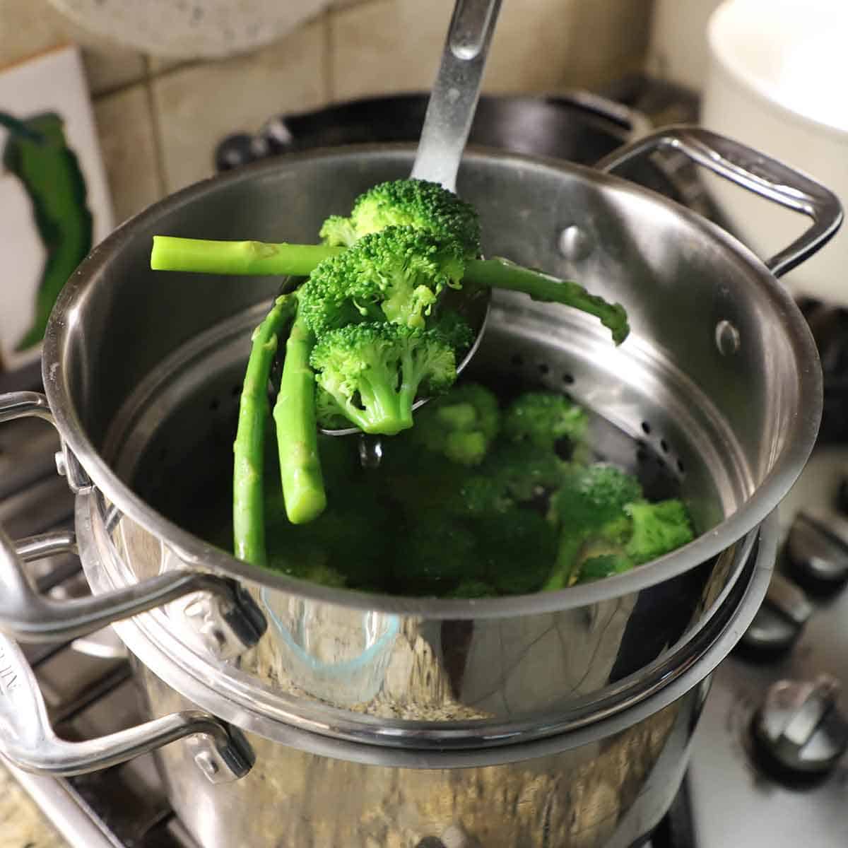 A person using a slotted spoon to remove fresh broccoli florets and asparagus spears from a pot of boiling water.