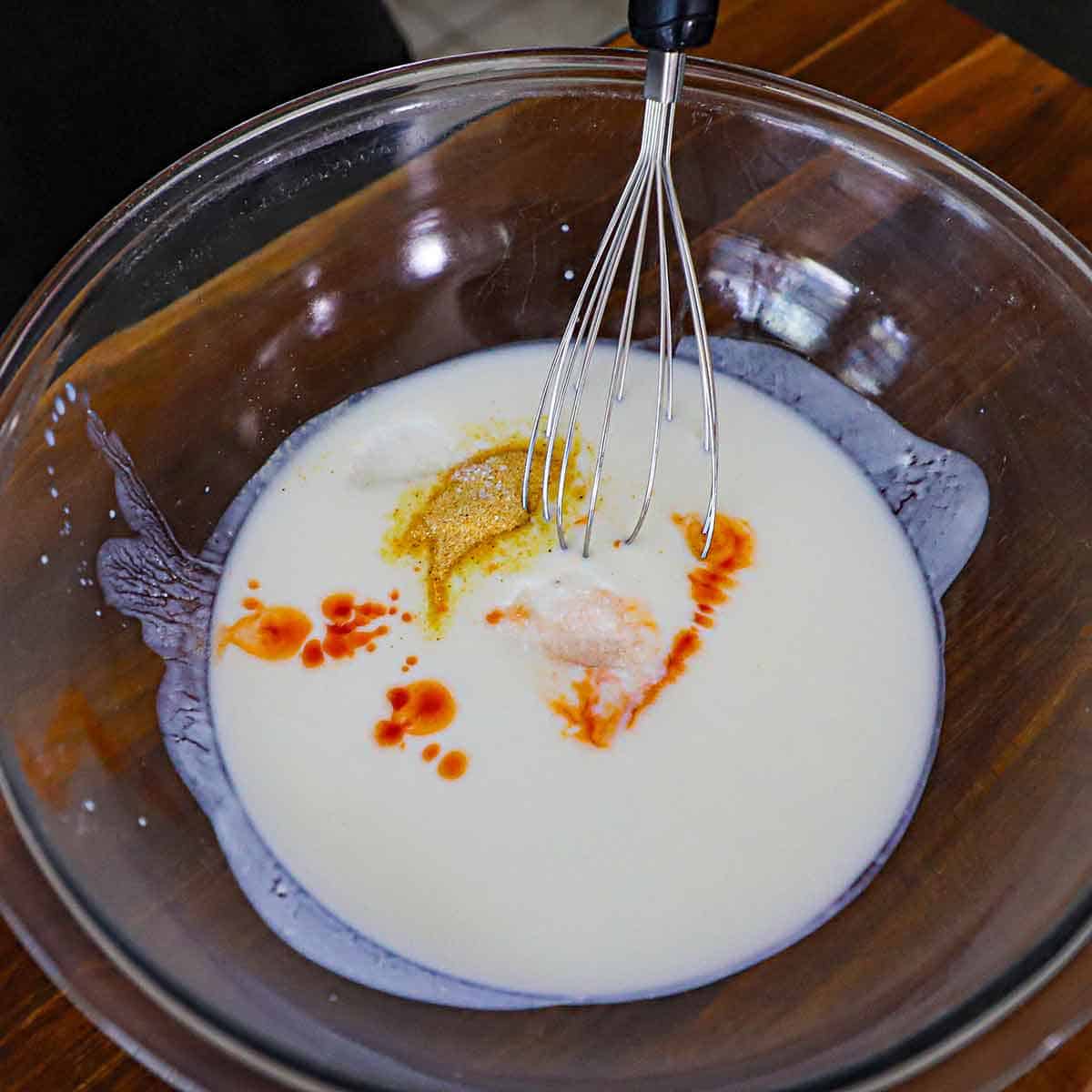 A person using a wire whisk to combine buttermilk with hot sauce and garlic powder in a large glass bowl.