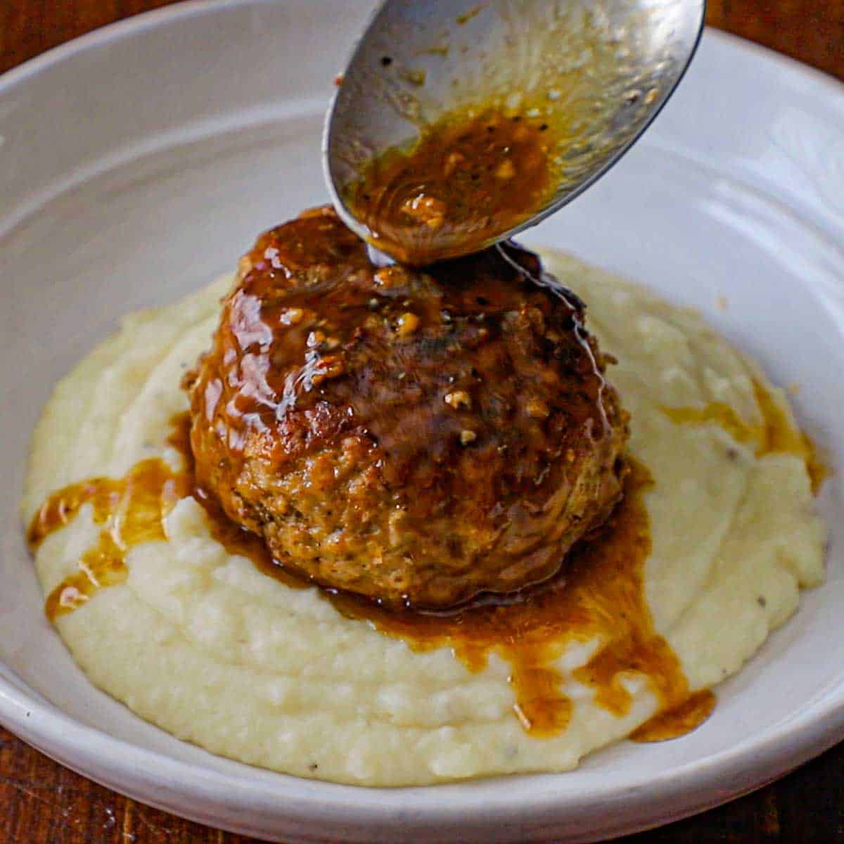 A large serving spoon being used to spoon a buttery beef au jus sauce over a large cooked Dutch meatball that is resting on a bed of mashed potatoes in a shallow white bowl.