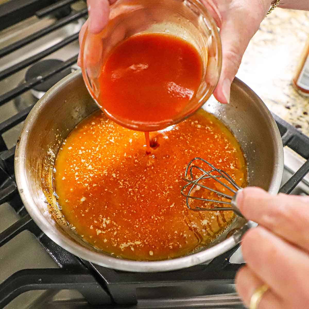 A person pouring Cayenne hot sauce from a small glass bowl into a small skillet filled with melted butter on a gas stove.