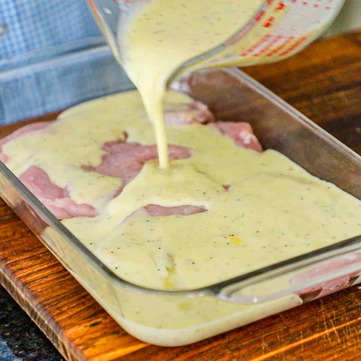 A person pouring seasoned buttermilk over two thinly pounded pork loin chops that are resting in a shallow glass baking dish.