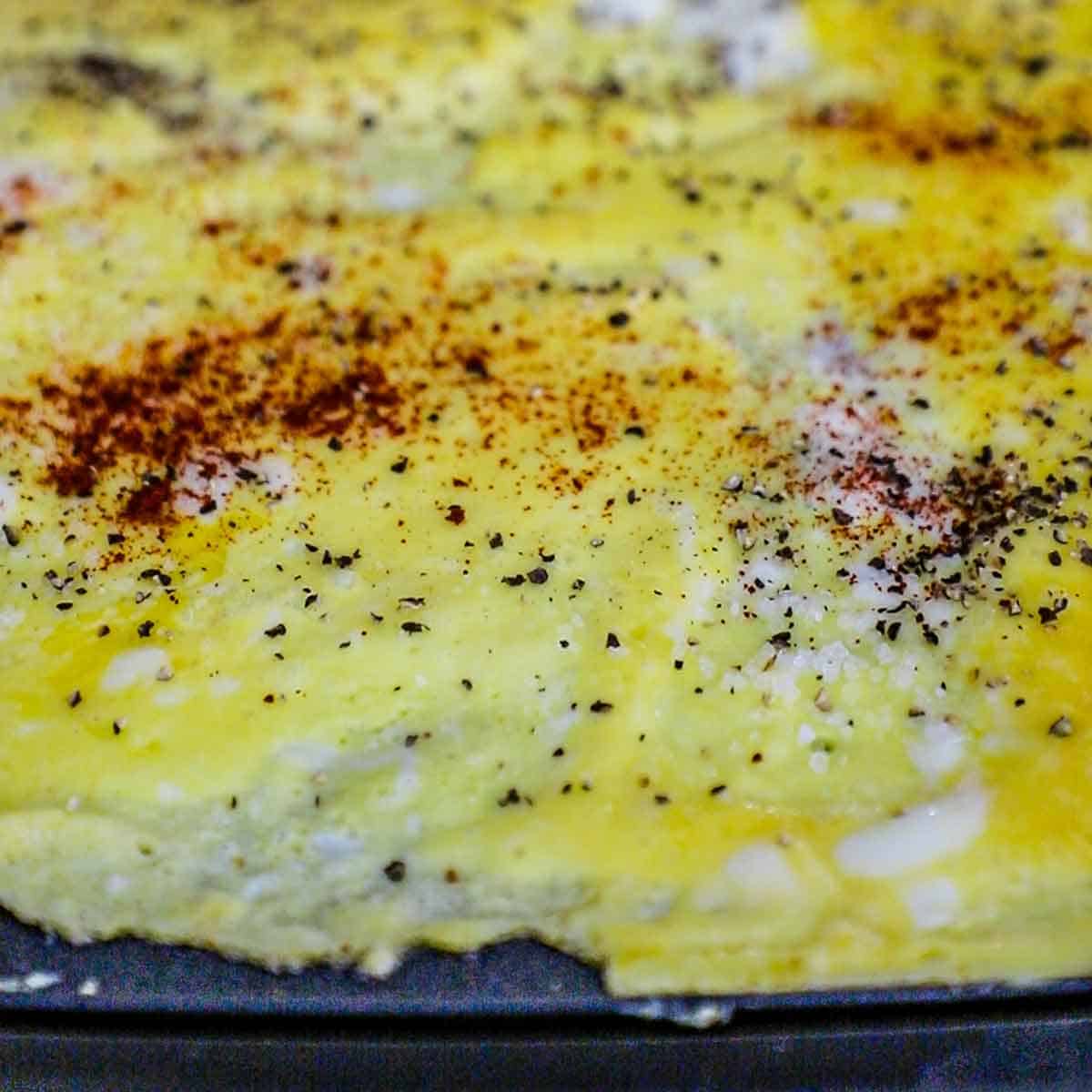 A close-up view of a thin layer of scrambled eggs that have been cooked and set on a griddle and seasoned with salt, pepper, and cayenne pepper.