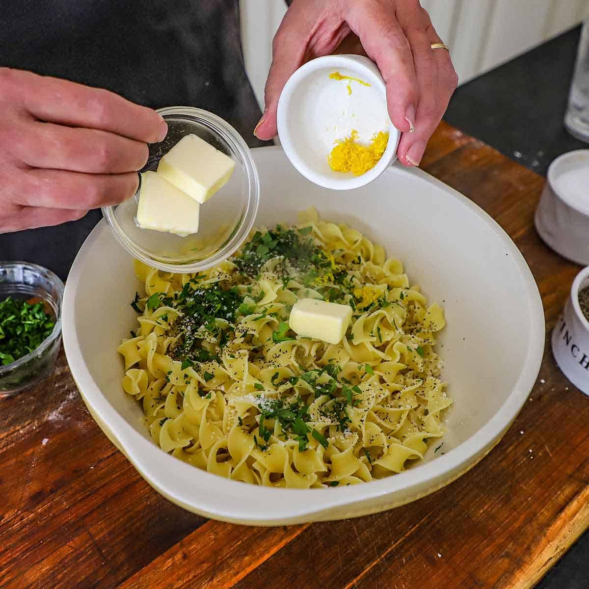 A person transferring pads of butter from a bowl in one hand and lemon zest from another small bowl in his other hand all into a large bowl filled with cooked egg noodles, herbs, and seasonings.