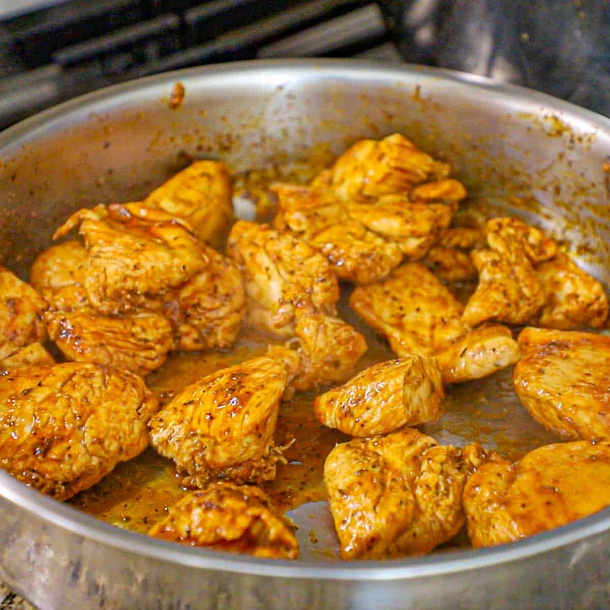 Large chunks of chicken that have being seasoned with homemade Cajun seasoning being seared in oil in a large silver skillet.