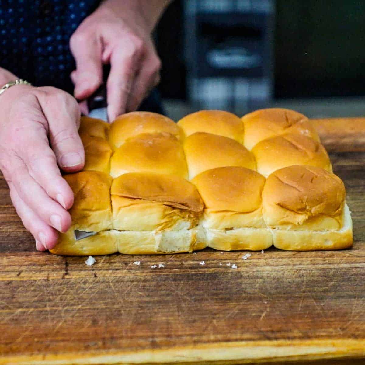 A person using a large serrated knife to cut through the middle of a flat of Hawaiian slider rolls on a wooden cutting board.