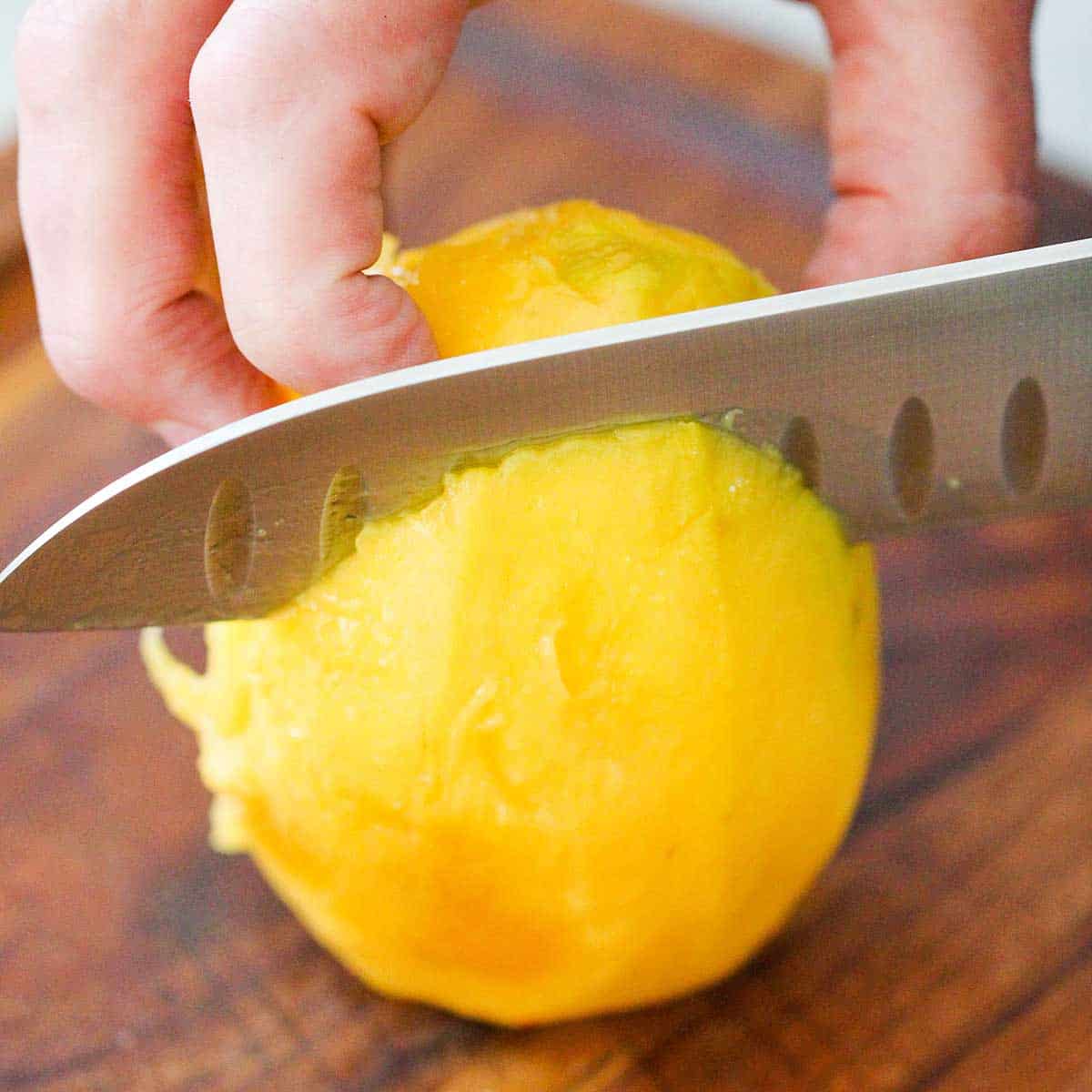 A person using a chef's knife to slice into a peeled, fresh mango on a wooden cutting board.