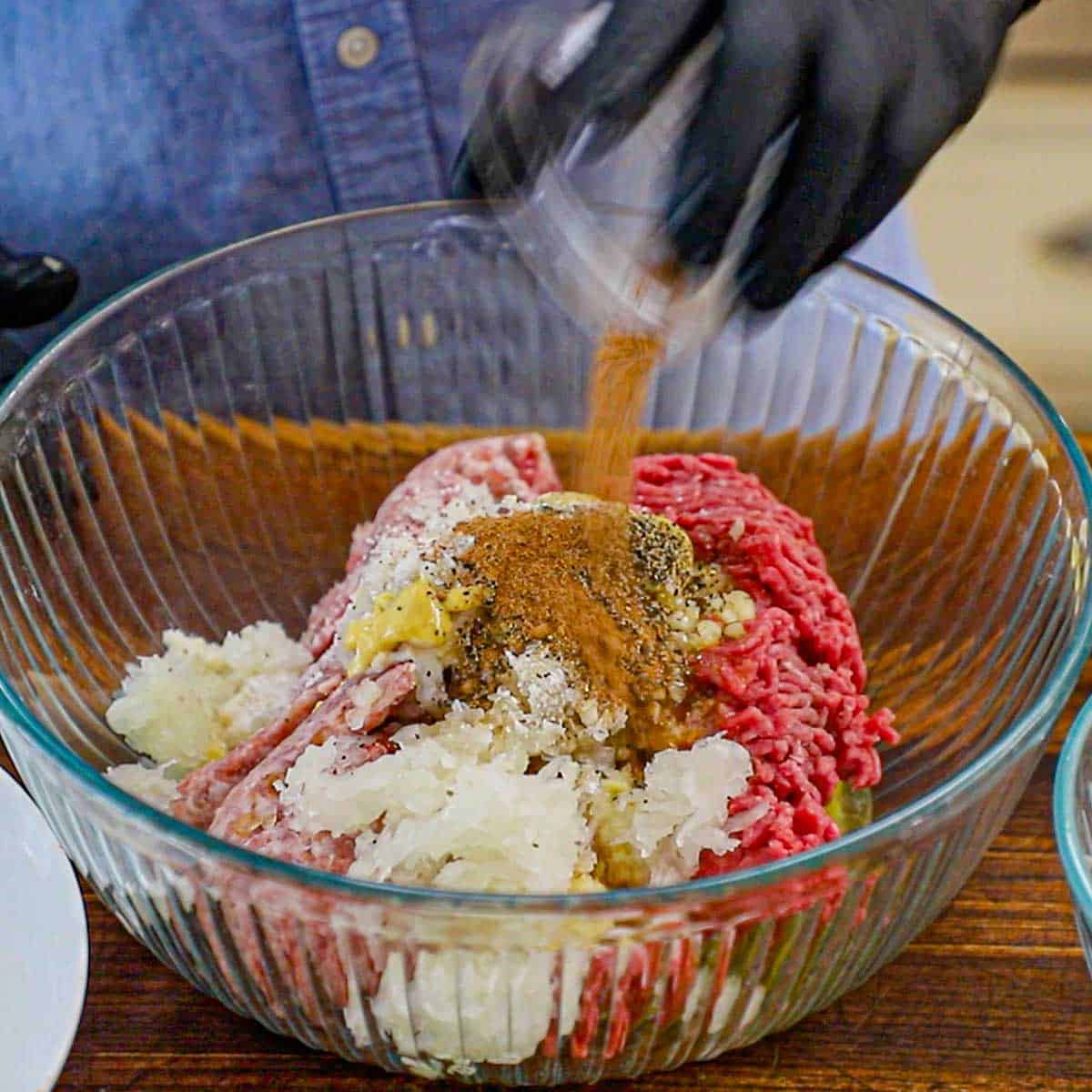 A person adding ground nutmeg from a small bowl into a larger glass bowl filled with ground meat, shredded onions, garlic, mustard, and seasonings.