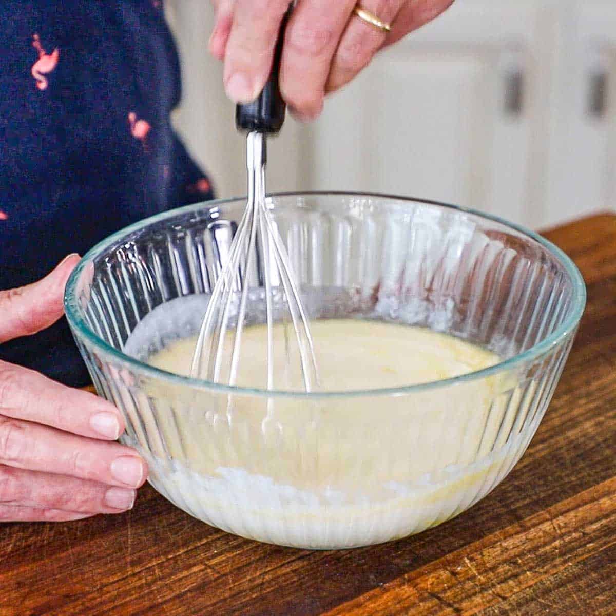 A person using a whisk to combine buttermilk with melted butter, eggs, and vanilla extract in a glass bowl on a wooden cutting board.