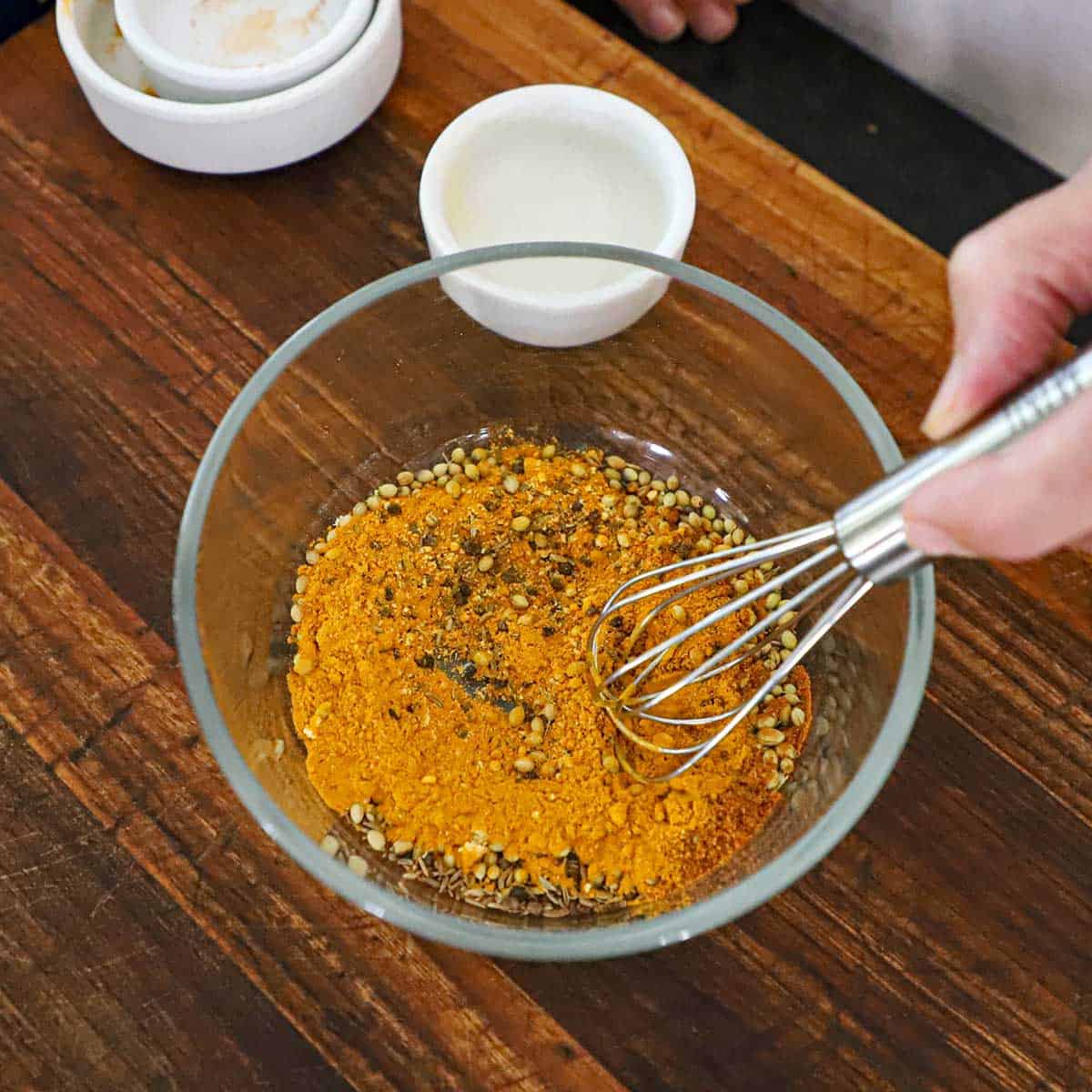 A person using a small whisk to combined toasted spices with cayenne pepper and ground mustard in a small glass bowl on a wooden cutting board.