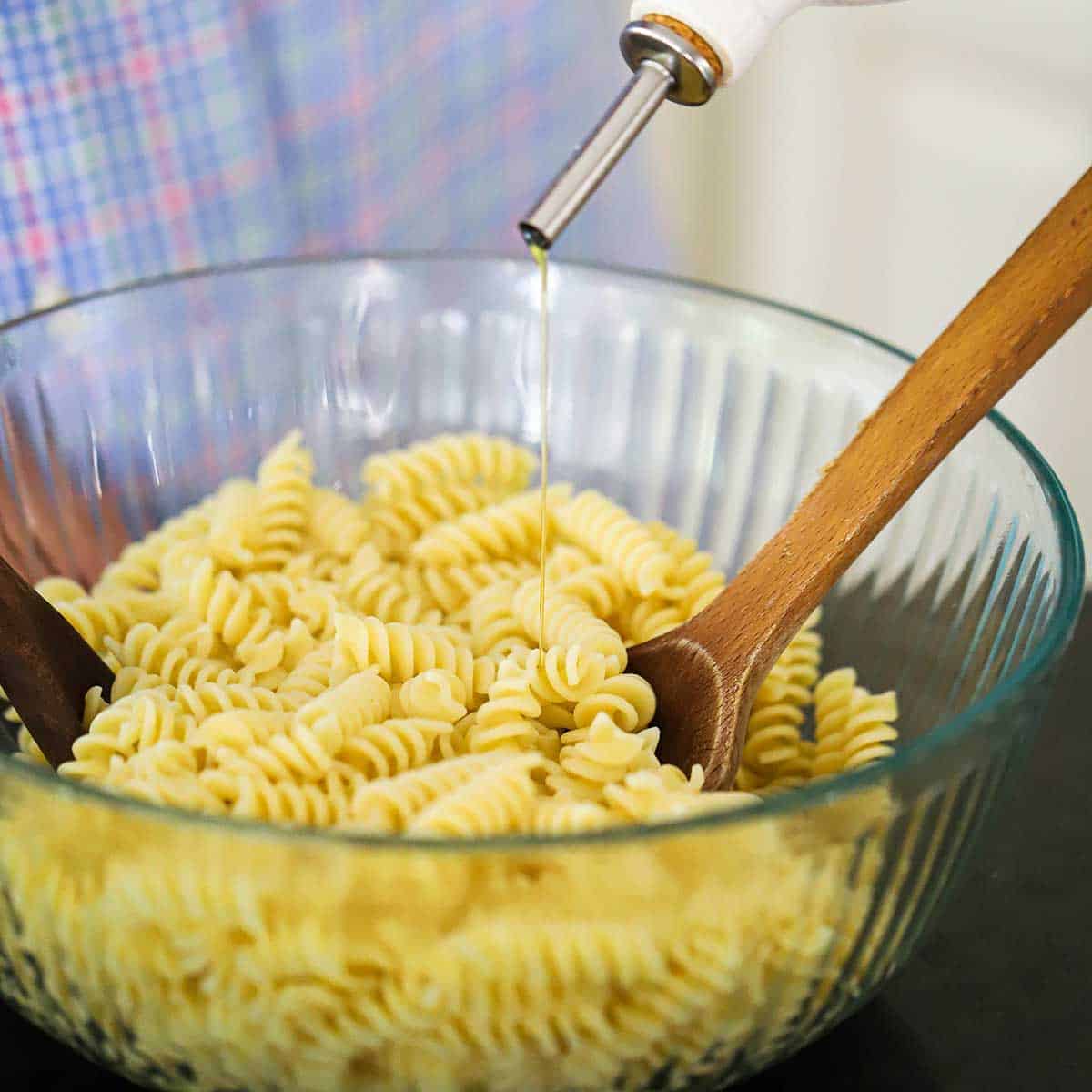 A person pouring olive oil from a oil dispenser into a glass bowl filled with cooked fusilli pasta.