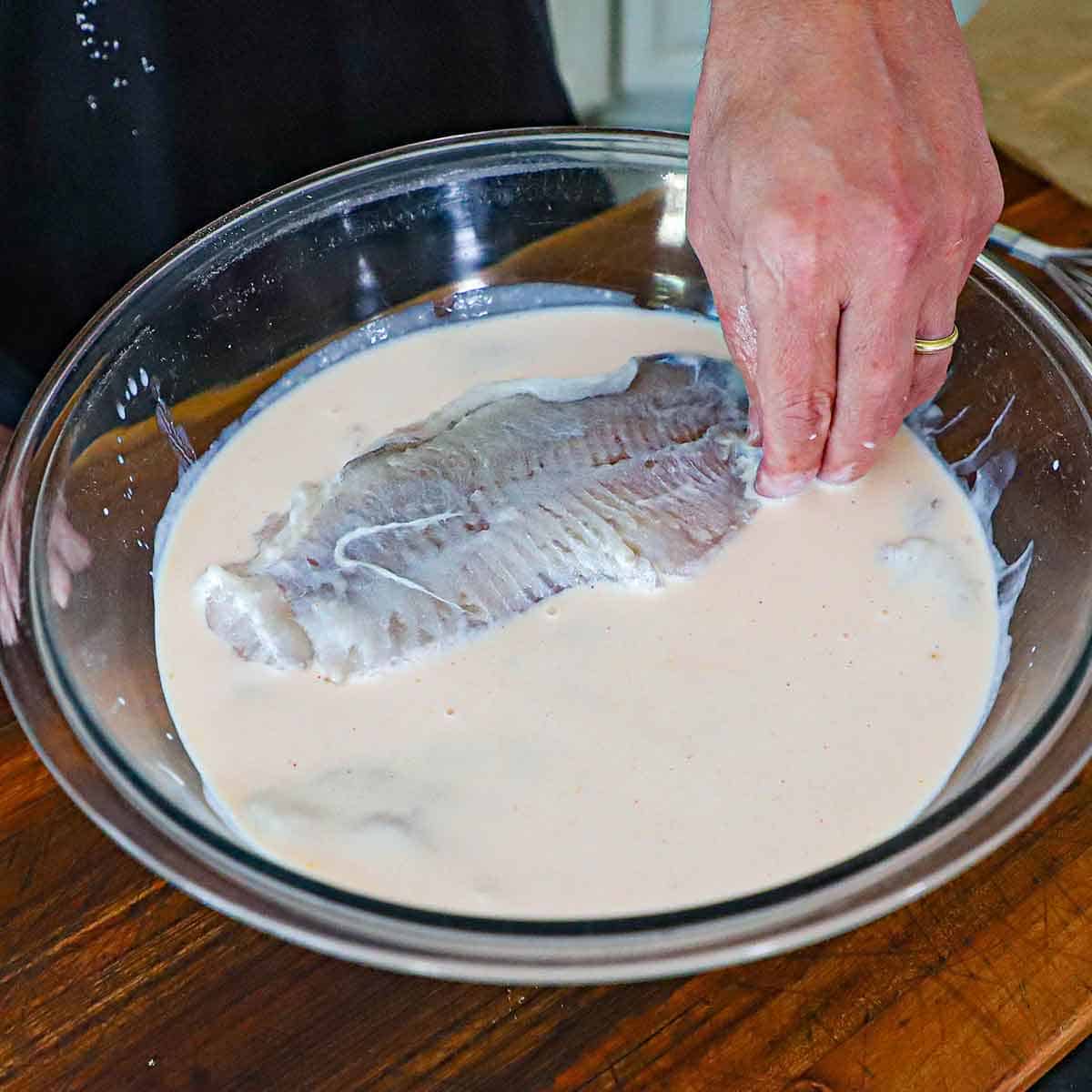 A person using his hand to submerge a fresh catfish filet in a large glass bowl filled with buttermilk combined with hot sauce and garlic powder.