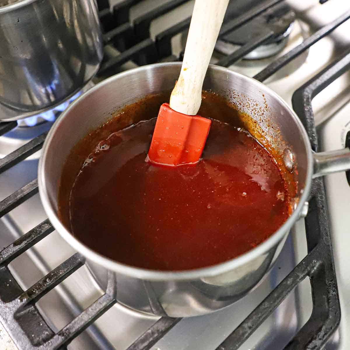 A person using a red spatula to stir a homemade honey barbecue sauce in a saucepan on a gas stove.