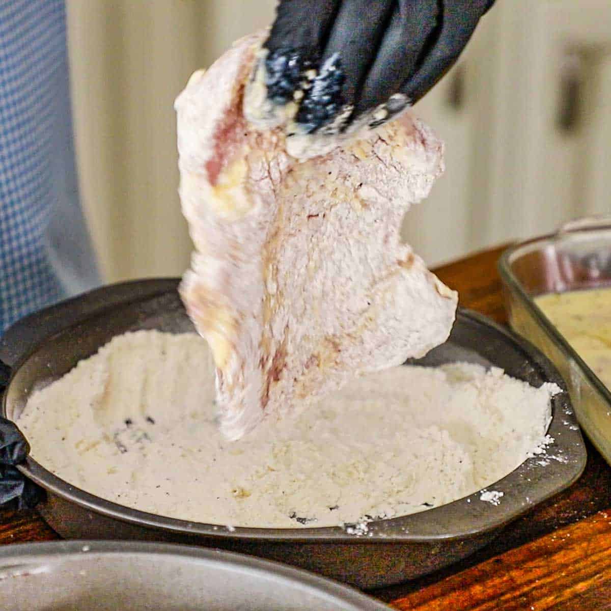 A person dredging a buttermilk-coated thin pork loin chop through seasoned flour in a circular baking pan.