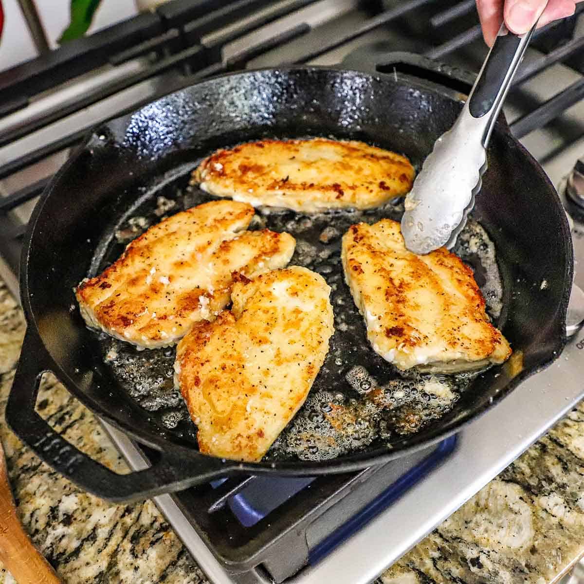 Person using metal tongs to flip floured and seasoned chicken breasts that are being seared in a large cast-iron skillet on a gas stove.
