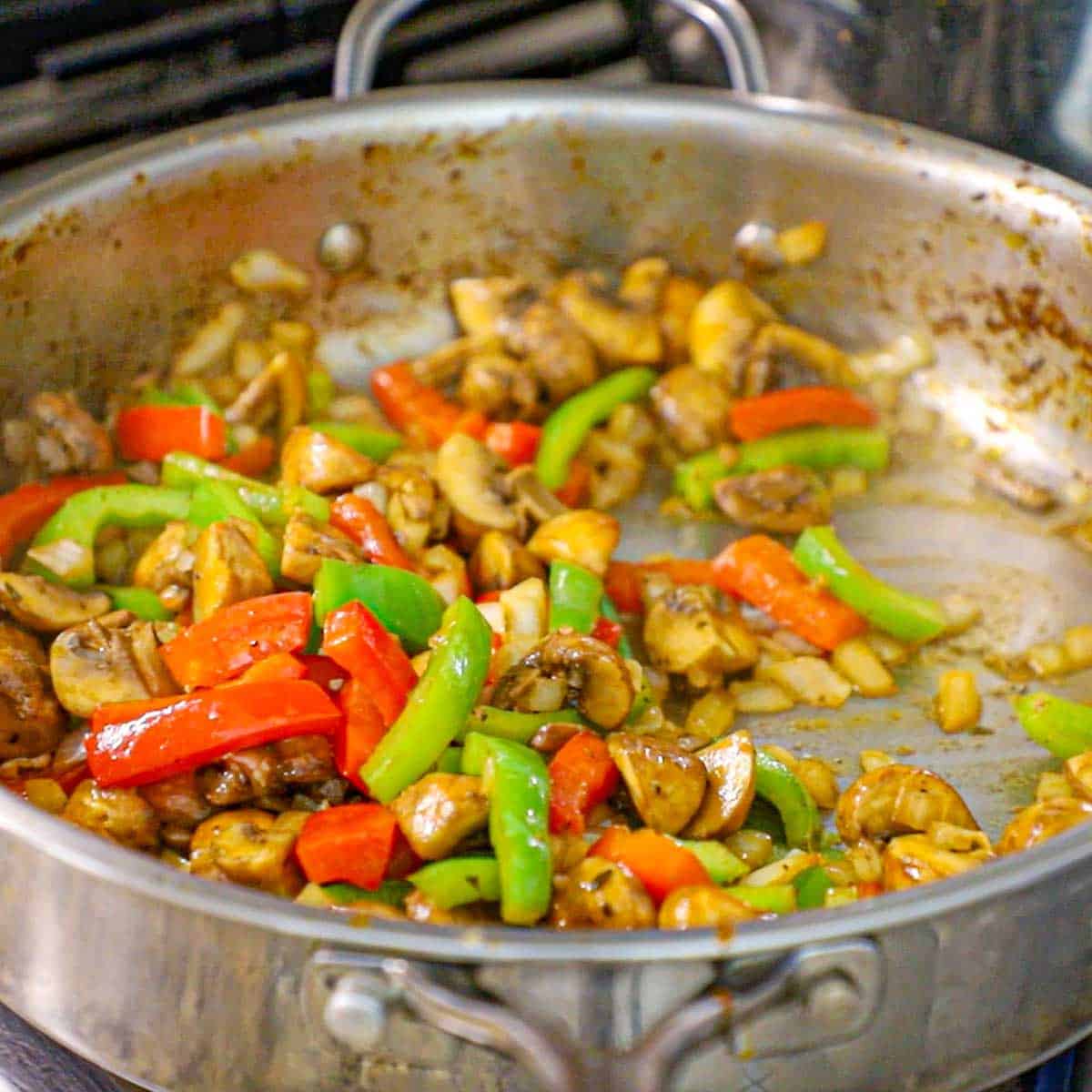 A straight-on view of a large stainless steel skillet filled with strips of red and green bell peppers, chopped onions, and quartered mushrooms that have been sautéed in olive oil.