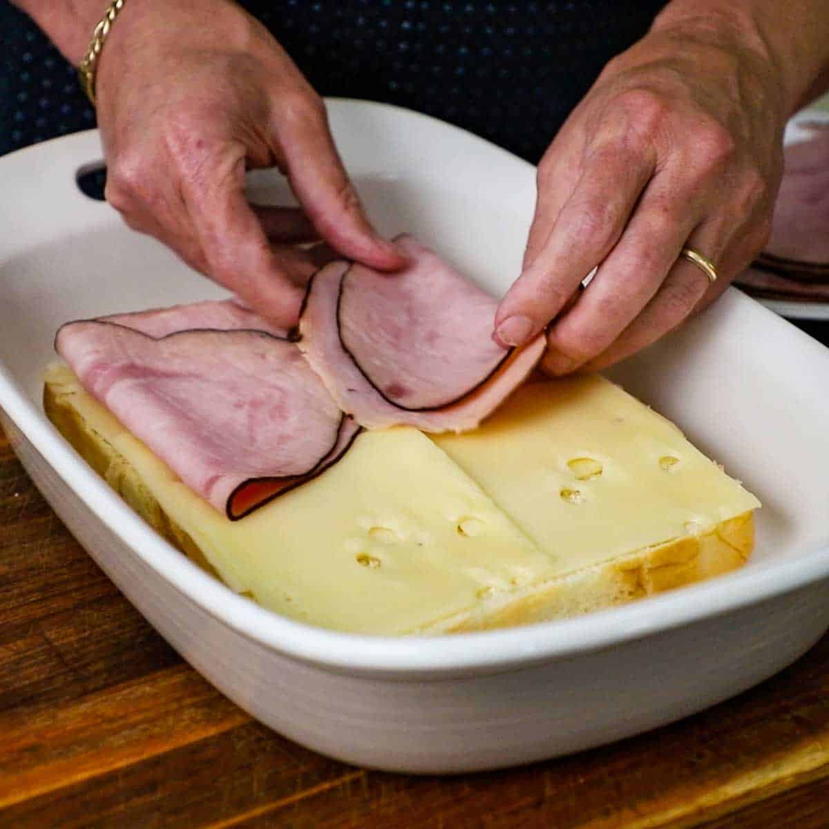 A person placing folded slices of black forest ham over slices of Swiss cheese that are resting on the bottom half layer of Hawaiian slider buns.