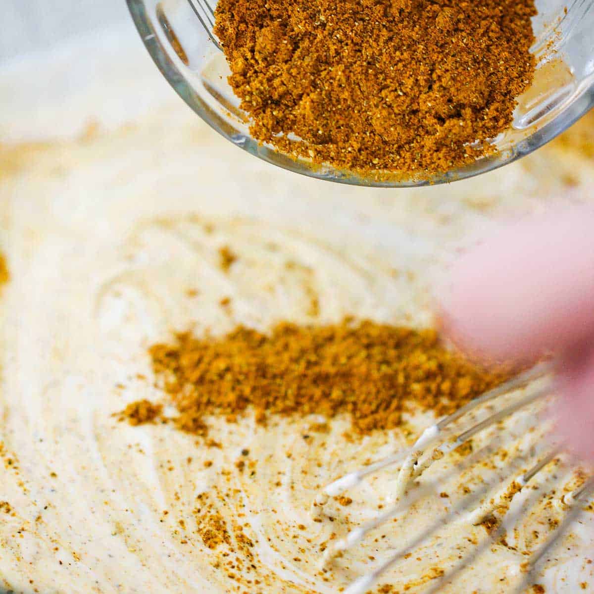 A person transferring homemade curry powder from a small glass bowl into a mayonnaise and yogurt dressing in a glass bowl.