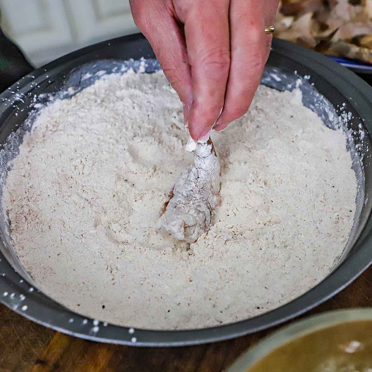 A person is holding a raw shrimp that has been butterflied by the tail and dredging through seasoned flour in a round baking pan.