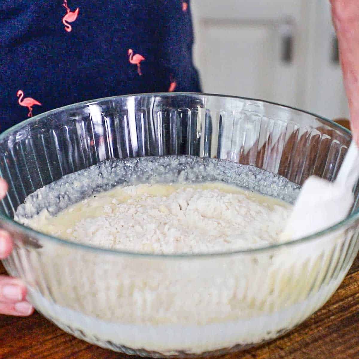 A person using a large white rubber spatula to combine the wet buttermilk pancake ingredients with the dry ingredients in a large glass bowl.