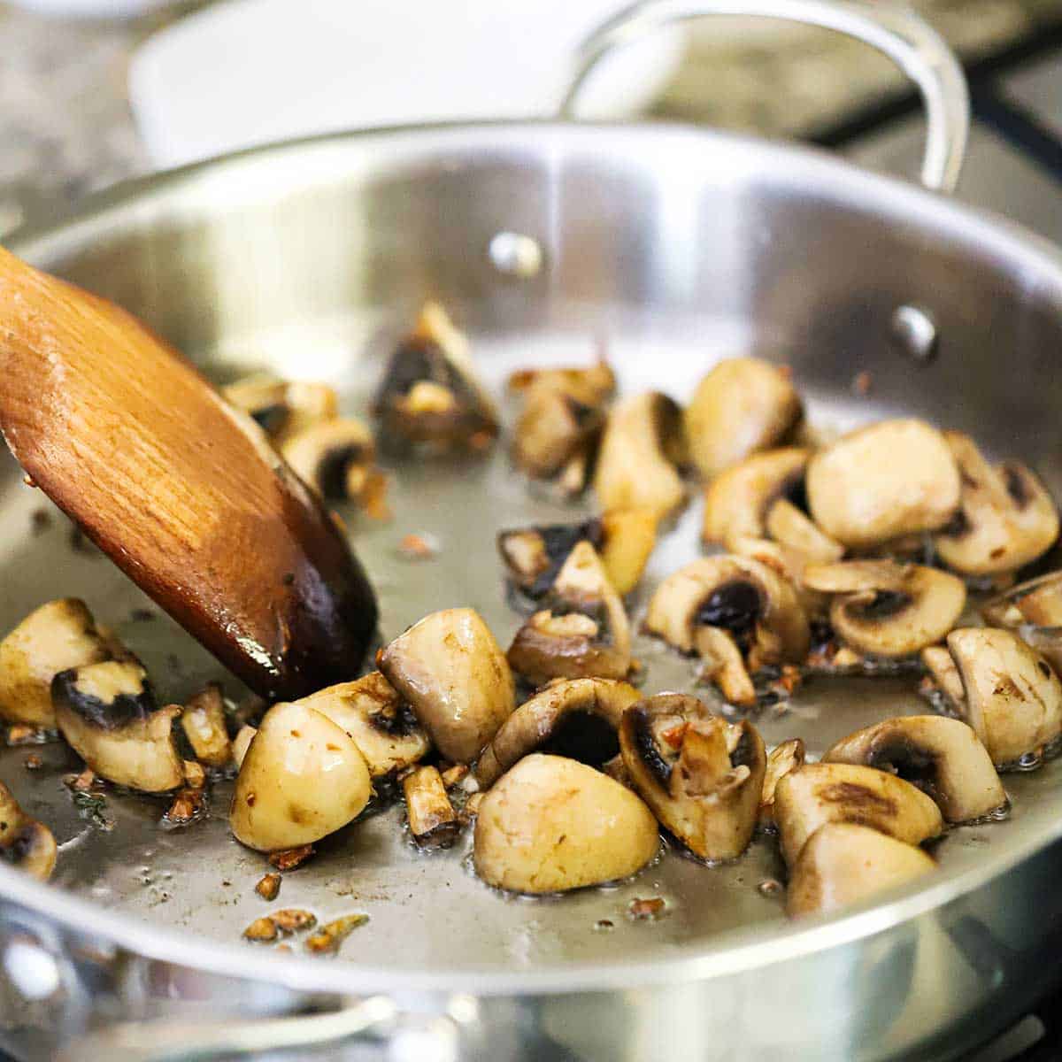 A person using a wooden spatula to stir quartered baby Bella mushrooms that are being sautéed in a large stainless steel skillet.