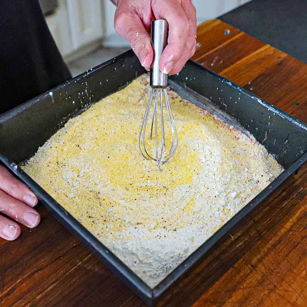 A person using a small whisk to combine cornmeal, flour, and seasonings in a square baking pan on a wooden cutting board.