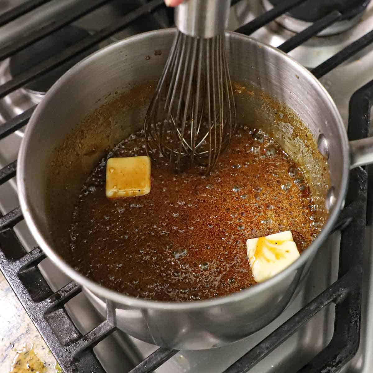 A person using a whisk to stir a simmering maple Bourbon glaze with two pieces of butter melting in the sauce.