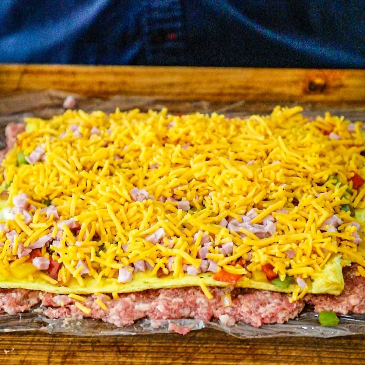 A large rectangular piece of plastic wrap that is topped with a layer of uncooked breakfast sausage, a layer of cooked eggs, sautéed peppers and onions, cubed ham, and shredded cheddar cheese, all on wooden cutting board.