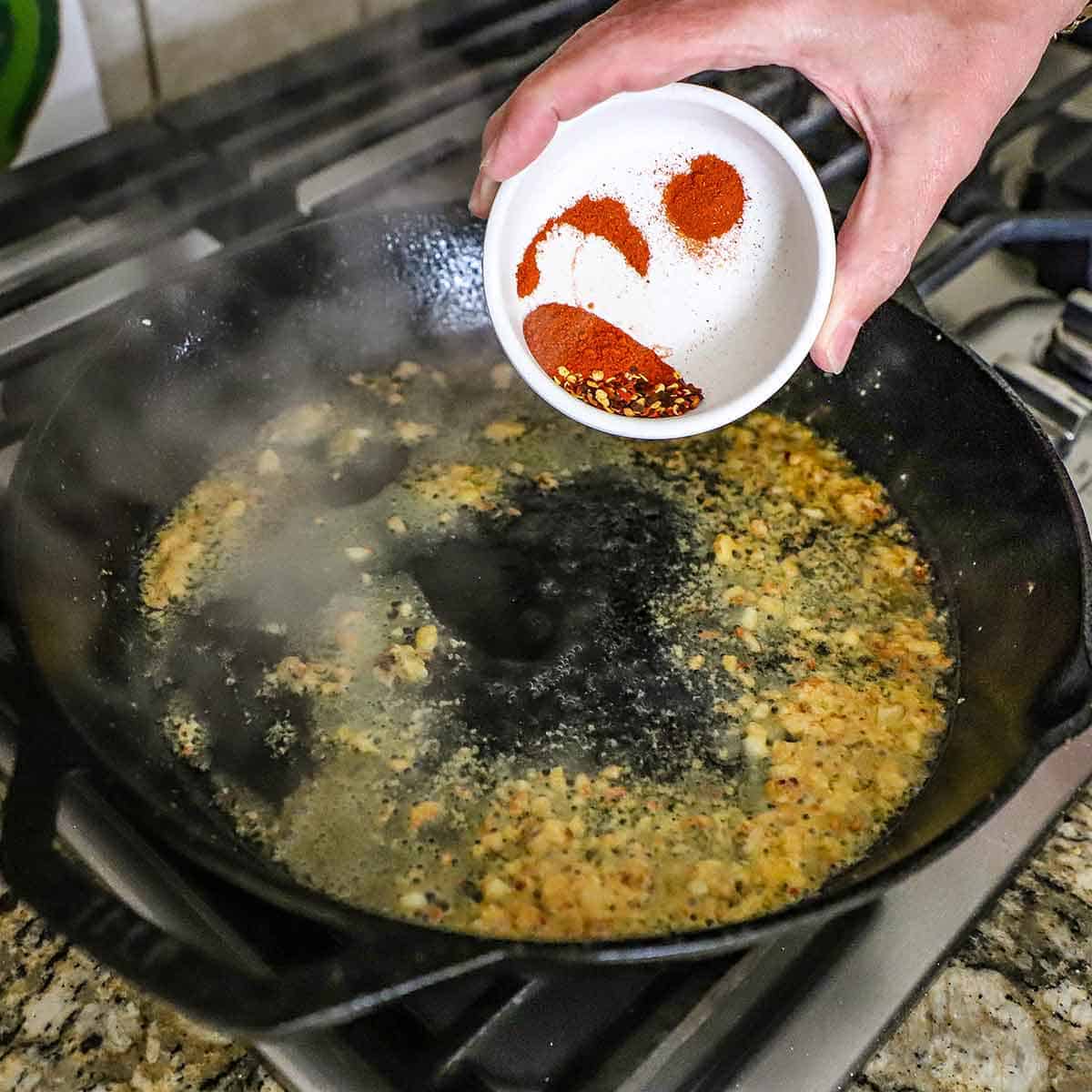 A person transferring spices from a small white bowl into a large cast-iron skillet that is filled with simmering minced garlic in oil and pasta water.