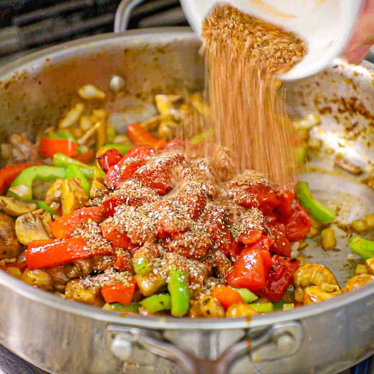 A person pouring homemade Cajun seasoning from a small white bowl into a large skillet filled sautéed bell peppers, onions, and mushrooms.
