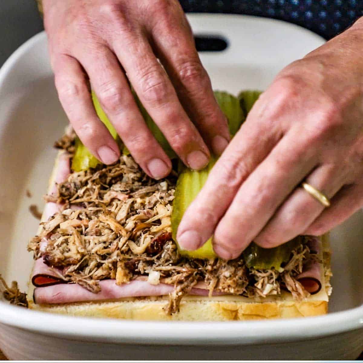 A person placing long dill pickle sandwiches planks on top of shredded seasoned pork, sliced ham, Swiss cheese slices, and the lower half of a rack of Hawaiian slider buns.