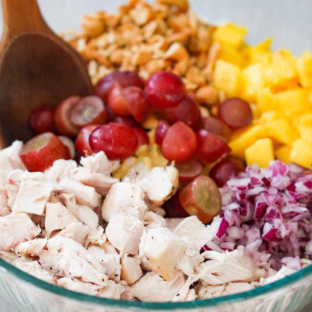 A bowl filled with diced cooked chicken, chopped red onion, diced fresh mango, roasted cashews, and sliced red grapes, with a wooden spoon inserted off to the side.