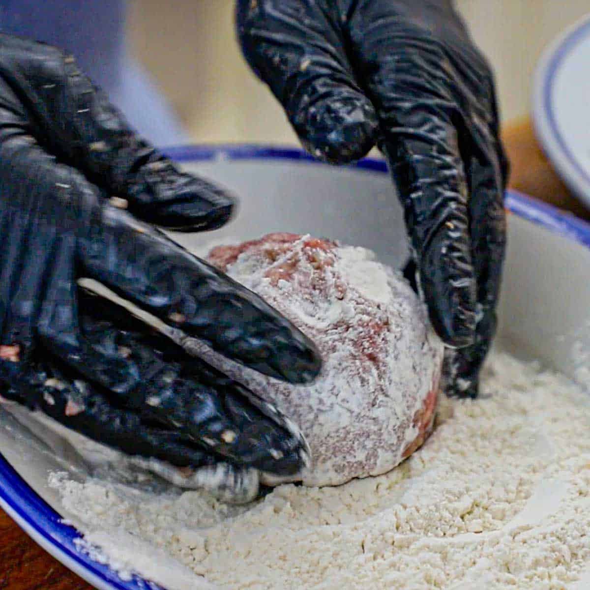 A person wearing black latex gloves gently rolling a large uncooked Dutch meatball in flour on an oval platter.