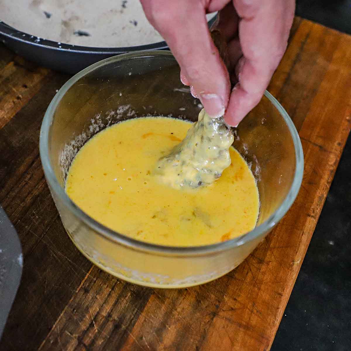 A person holding a raw butterflied shrimp in an egg and buttermilk mixture in a small glass bowl on a wooden cutting board.