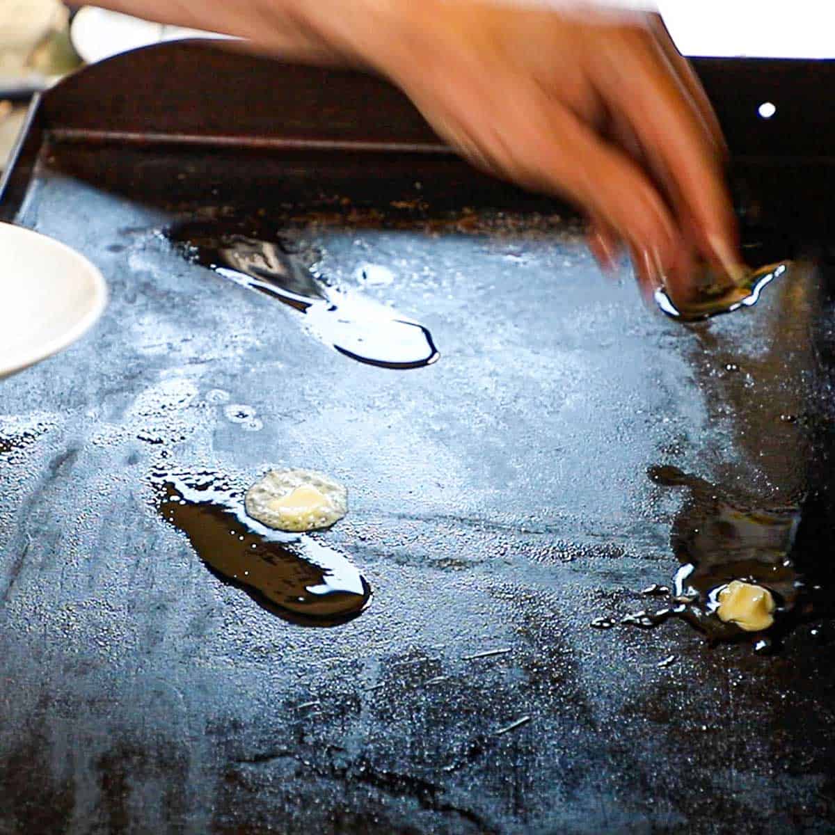 A person dropping a small cube of butter onto a small pool of oil that is simmering on a large outdoor griddle.